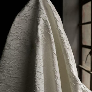 Könnte beinhalten: Off-white Stoff mit einer strukturierten, gewellten Oberfläche. Der Stoff ist drapiert und erzeugt weiche Falten und Schatten. Das Material scheint dick und undurchsichtig zu sein, mit einem dezenten Glanz. Der Hintergrund ist dunkel, mit einem Fenster rechts.