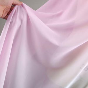 May include: A close-up of a sheer, light pink fabric with soft folds and subtle gradients of color. The fabric is held up, creating gentle waves and shadows. The material appears delicate and translucent, suggesting a lightweight textile.