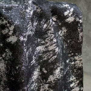 May include: Close-up of a textured fabric with a black and silver abstract pattern. The fabric has a rough, uneven surface with a mix of dark and light tones, creating a dynamic visual effect. The design appears to be a blend of brushstrokes and speckled elements.