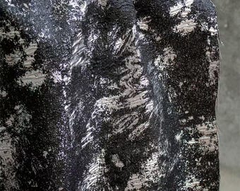 May include: Close-up of a textured fabric with a black and silver abstract pattern. The fabric has a rough, uneven surface with a mix of dark and light tones, creating a dynamic visual effect. The design appears to be a blend of brushstrokes and speckled elements.