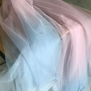 Puede incluir: Primer plano de tela transparente en degradado de colores. La tela transiciona de un azul claro a un tono rosa suave. El material parece delicado y fluido, con pliegues y capas visibles.