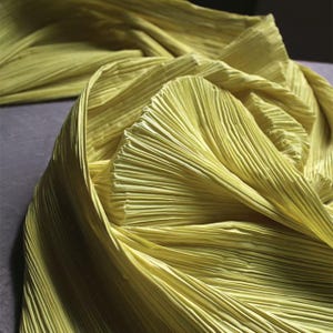May include: A close-up shot of a folded, pleated fabric in a pale yellow-green colour. The fabric has a textured, ribbed appearance with vertical lines. The folds create a layered effect, with shadows adding depth. The overall aesthetic is soft and flowing.
