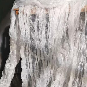 Könnte beinhalten: Nahaufnahme eines weißen, fließenden Stoffes mit welliger Textur. Der Stoff ist über eine Holzstange drapiert und erzeugt vertikale Linien und ein Gefühl von Bewegung. Das Material wirkt leicht und durchscheinend.