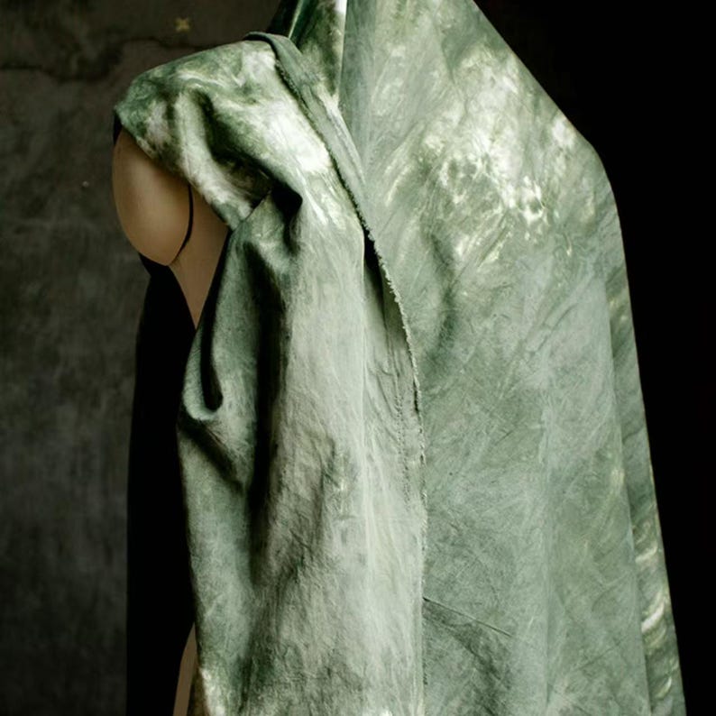 Puede incluir: Una tela drapeada en tonos verdes y blancos, posiblemente lino o seda, se exhibe sobre un maniqu&iacute;. La tela tiene un efecto tie-dye o acuarela, con un patr&oacute;n moteado. La est&eacute;tica general es natural y org&aacute;nica.