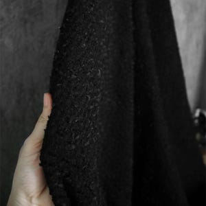 May include: Close-up of a black fabric with a textured, nubby surface. The fabric is held in a hand, revealing its depth and the play of light and shadow across its surface. The material appears to be thick and possibly woven.