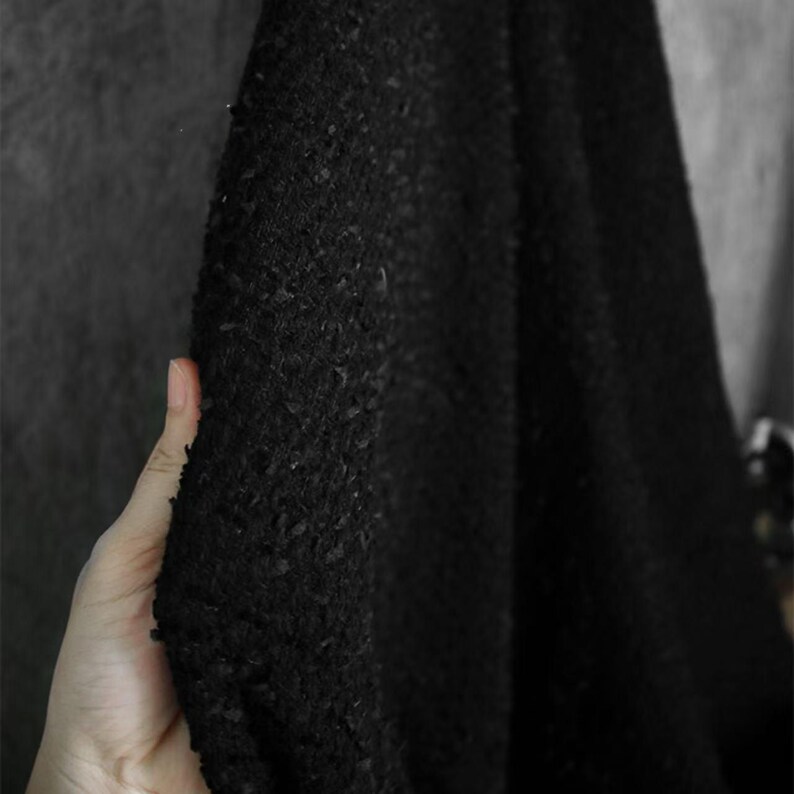 May include: Close-up of a black fabric with a textured, nubby surface. The fabric is held in a hand, revealing its depth and the play of light and shadow across its surface. The material appears to be thick and possibly woven.