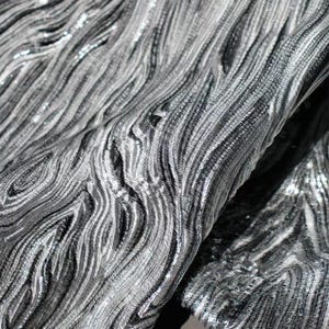 Puede incluir: Primer plano de una tela plateada con una superficie texturizada y ondulada. La tela tiene un brillo metálico, con la luz reflejándose en las crestas y creando un patrón dinámico. El material parece estar doblado, mostrando profundidad y dimensión.