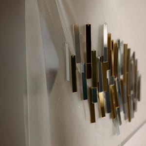 May include: Abstract wall art composed of rectangular metal pieces in silver, gold, and black. The pieces are arranged in an overlapping pattern, creating a modern, geometric design. The artwork is mounted on a white surface, with a sheer curtain visible.