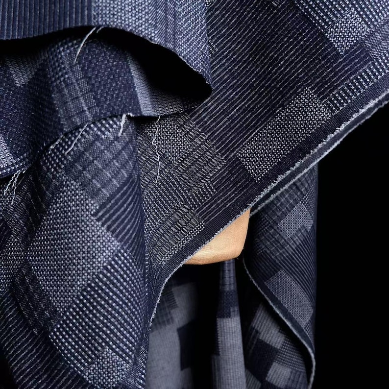 May include: A dark blue fabric featuring a geometric pattern of squares and rectangles with varying textures. The design consists of alternating blocks of fine horizontal lines, vertical stripes, and dotted grids, creating a complex, layered visual effect.