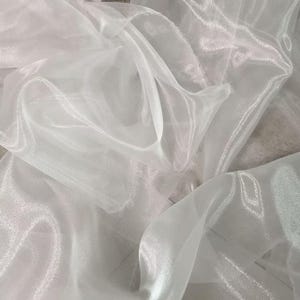 May include: A close-up of white, sheer fabric with a shimmering, translucent quality. The fabric is draped and folded, creating soft shadows and highlights. The material appears lightweight and flowing, suitable for apparel or decorative purposes.