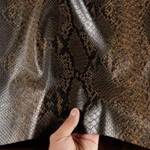 May include: Close-up of a snakeskin pattern in shades of brown, tan, and gray. The textured surface mimics the scales of a snake. The material appears to be flexible and possibly leather or a similar fabric.