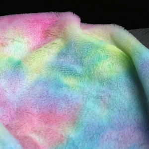 Op de afbeelding: Close-up van een zachte, donzige deken met een tie-dye patroon. De deken heeft een mix van levendige kleuren, waaronder roze, geel, groen en blauw. Het pluche materiaal heeft een getextureerde uitstraling.