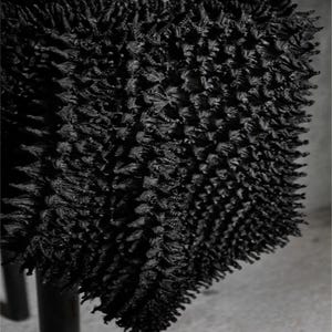 May include: Close-up of a black, textured textile. The fabric has a dense, three-dimensional surface with a multitude of small, irregular protrusions. The material appears to be soft and possibly woven or knitted, with a dark, matte finish. The textile is draped over a dark support structure.