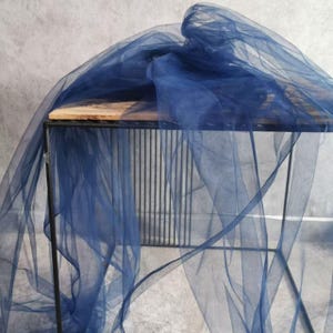 May include: Navy blue sheer fabric draped over a black metal table. The fabric is lightweight and translucent, with a delicate texture. The table has a wooden top and a geometric metal frame. The fabric is bunched up in places, creating soft folds and shadows.