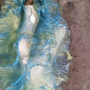 Puede incluir: Una tela fluida e iridiscente en tonos azules y verdes, con bordes con volantes. La tela está drapeada sobre un maniquí, creando un efecto de capas. El material tiene una calidad brillante, que capta la luz y crea un efecto visual dinámico.
