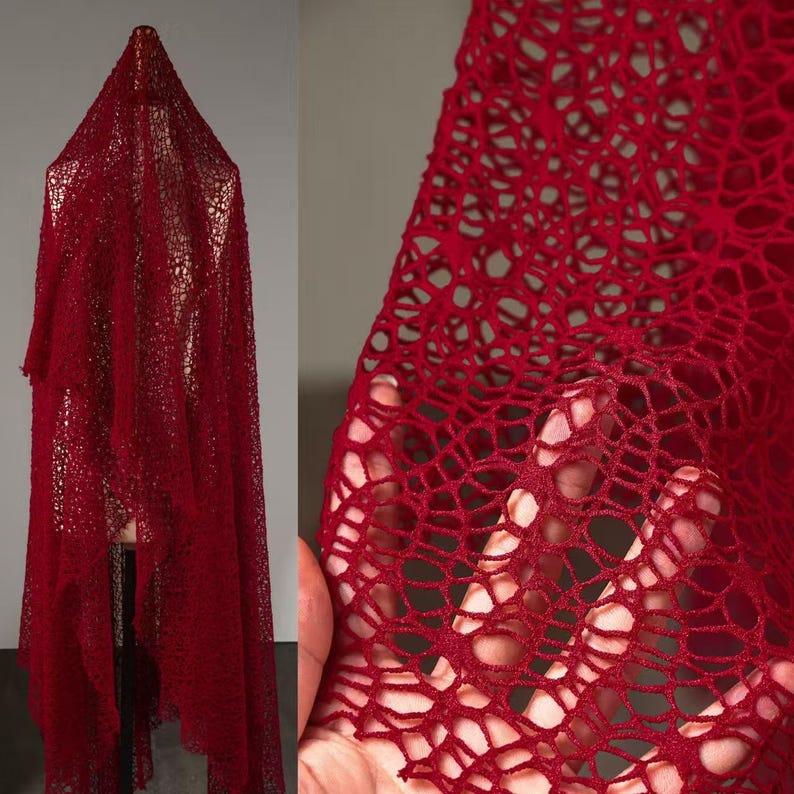 May include: A deep red, open-weave fabric, possibly lace or netting, is draped over a form. The fabric's texture is visible in a close-up shot, revealing a web-like pattern. The material appears lightweight and airy, with a rich, saturated color.