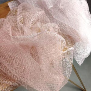May include: A sheer, lightweight fabric with a delicate, open-weave mesh structure. The material features a soft, pale pink hue that transitions into a lighter, translucent white section, creating a layered, ethereal appearance with a subtle sheen.