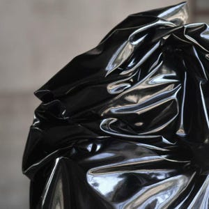 Op de afbeelding: Close-up van een glanzend, zwart materiaal met een reflecterend oppervlak. De stof is sterk gekreukt en gevouwen, waardoor schaduwen en highlights ontstaan. Het algehele effect is strak en modern.