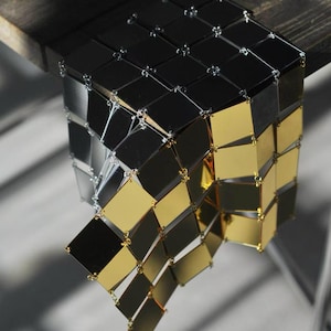 May include: A geometric, cube-shaped decorative object composed of connected, reflective square tiles. The tiles alternate between black, silver, and gold, creating a striking visual contrast. The object hangs from a wooden surface.