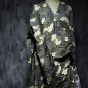 May include: A wrap-style garment featuring a layered, textured camouflage pattern in shades of dark gray, olive green, and cream. The fabric displays frayed, overlapping edges throughout the surface, creating a dimensional effect with a V-neckline.
