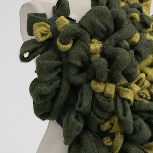 May include: A close-up of a textile art piece featuring looped and knotted fabric in shades of olive green and chartreuse. The fabric is arranged in a dense, textured pattern, creating a three-dimensional effect. The piece is mounted on a mannequin.