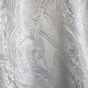 K&ouml;nnte beinhalten: Nahaufnahme eines silbernen Stoffes mit strukturiertem, marmoriertem Aussehen. Der Stoff hat ein wirbelndes Muster mit erhabenen, metallisch aussehenden Details. Der Gesamteffekt ist elegant und luxuri&ouml;s, geeignet f&uuml;r Kleidung oder Heimdekoration.