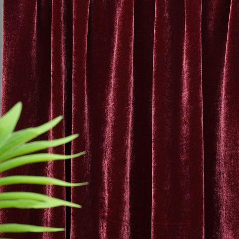 May include: Close-up of deep burgundy velvet curtains with vertical folds. The fabric has a soft, luxurious sheen. A few green plant leaves are visible in the lower left corner.