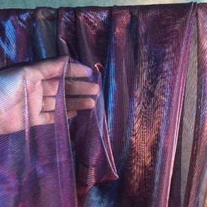 May include: A shimmering fabric in purple, blue, and pink hues. The material has a ribbed texture and is partially held up, showing its transparency. The light catches the fabric, producing a radiant effect. Ideal for clothing or decorative purposes.
