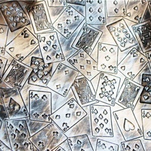 Op de afbeelding: Een metalen oppervlak met een herhalend reliëfpatroon van diverse speelkaarten, waaronder plaatjes en genummerde kaarten met harten, ruiten, klaveren en schoppen symbolen, afgewerkt in een verweerde, antiek ogende zilverkleur.