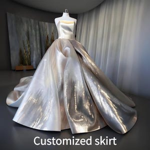 Puede incluir: Un vestido de baile sin tirantes de color marfil con una falda brillante y metálica. El corpiño es de color marfil liso y la falda fluye hacia una cola dramática. El texto "Customized skirt" está en la parte inferior de la imagen.