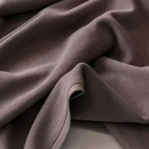 May include: Close-up of a folded, taupe fleece fabric. The material has a soft, textured surface with visible folds and creases. The edges are neatly finished, suggesting a blanket or throw.