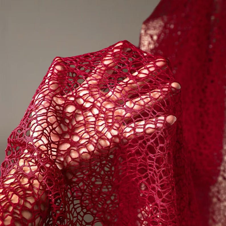 May include: Close-up of a deep red, open-weave fabric. The material is held up, revealing the intricate, interconnected pattern of the weave. The fabric appears soft and slightly sheer, with light passing through the holes.