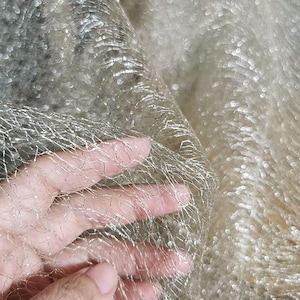 Pode incluir: Close-up de um tecido brilhante e texturizado com aparência de rede. O tecido é de cor dourada clara com fios prateados entrelaçados, criando um efeito cintilante. O material é macio e flexível, com um fundo ligeiramente desfocado.