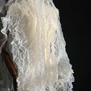 May include: A white, sheer fabric with a ruffled edge is draped over a mannequin. The fabric has a crinkled texture and appears lightweight. The dark background highlights the delicate, translucent material.