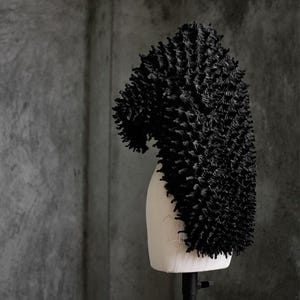 May include: A black, textured headpiece with a unique, spiky design. The head covering is displayed on a mannequin head against a gray background. The headpiece has a curved shape and a dense, three-dimensional texture.
