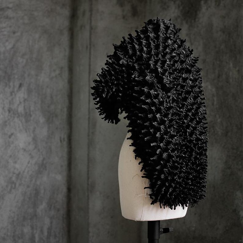 May include: A black, textured headpiece with a unique, spiky design. The head covering is displayed on a mannequin head against a gray background. The headpiece has a curved shape and a dense, three-dimensional texture.
