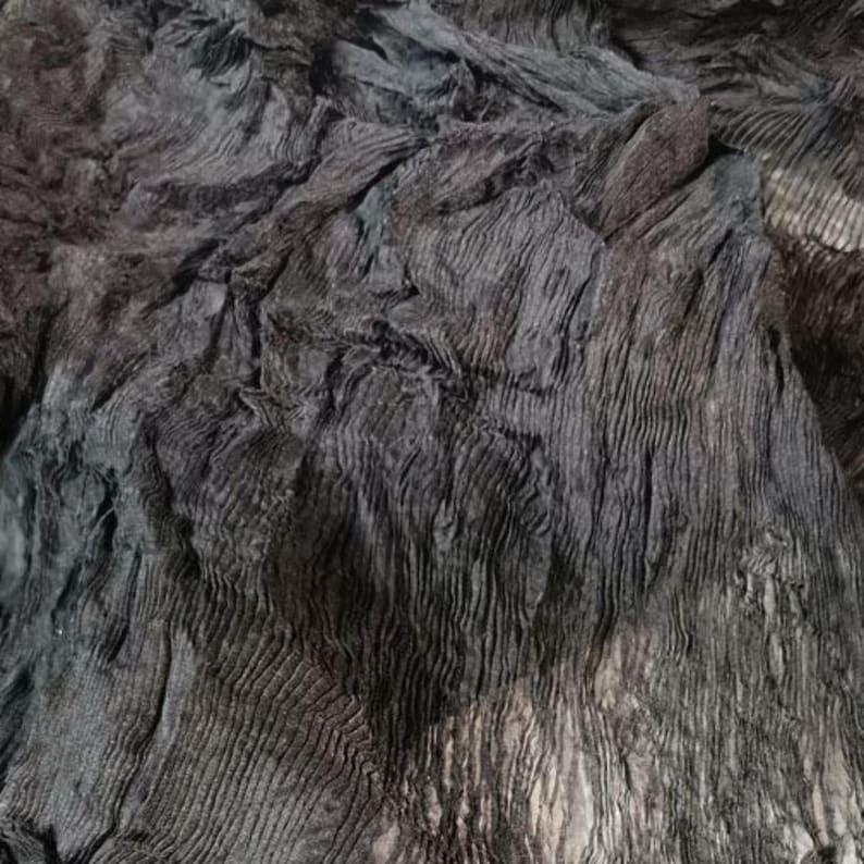 May include: Close-up of a dark gray fabric with a textured, wrinkled appearance. The fabric has a layered effect with visible ridges and folds, creating a sense of depth and dimension. The overall tone is muted, with subtle variations in shade.