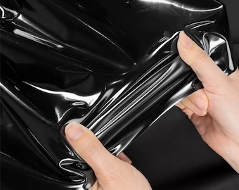 Puede incluir: Primer plano de una lámina de material negro brillante sostenida por dos manos. El material tiene una superficie reflectante con pliegues y arrugas visibles. La apariencia sugiere una tela flexible o similar.