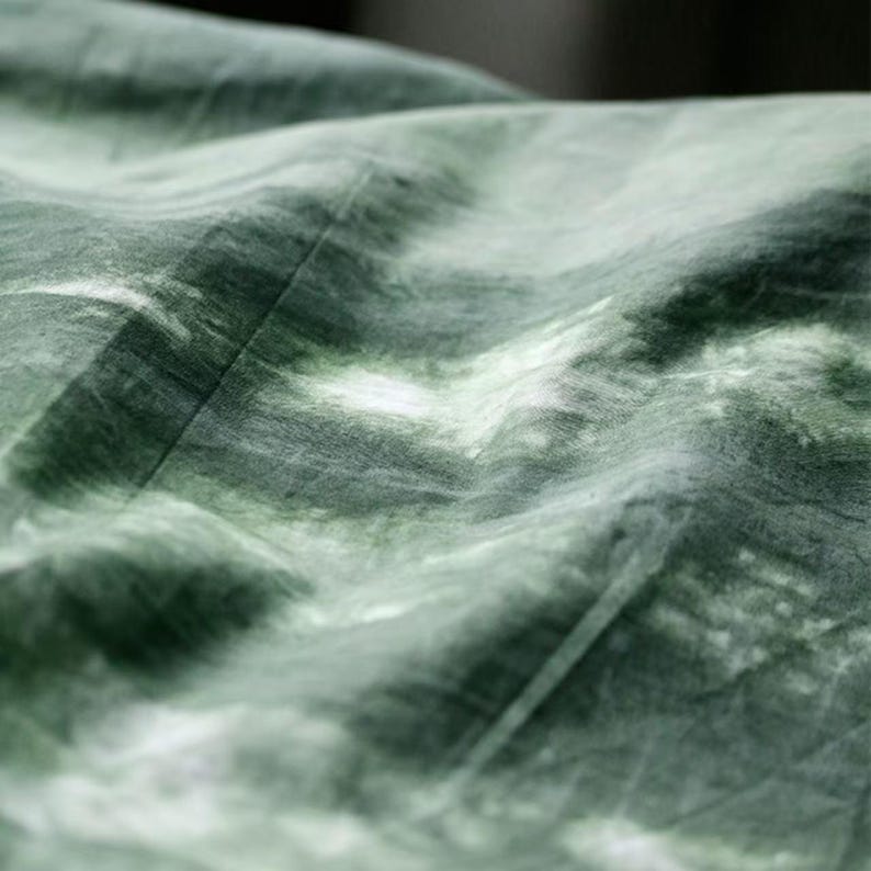 Puede incluir: Primer plano de una tela con un patr&oacute;n tie-dye en tonos verdes y blancos. La tela parece suave y fluida, con pliegues y texturas visibles. El efecto de tinte crea una apariencia marmolada.
