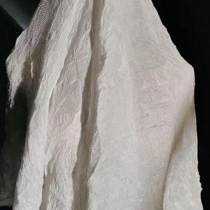 May include: A close-up of a white, textured fabric with a rippled pattern. The fabric is draped, creating soft folds and shadows. The material appears lightweight and semi-transparent, with a subtle sheen.