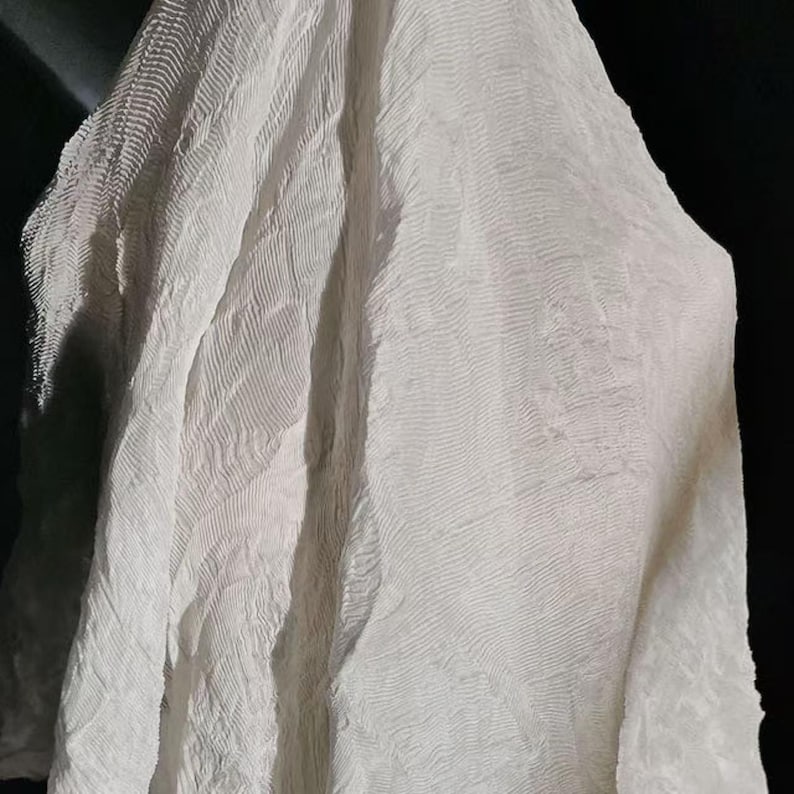 May include: A close-up of a white, textured fabric with a rippled pattern. The fabric is draped, creating soft folds and shadows. The material appears lightweight and semi-transparent, with a subtle sheen.