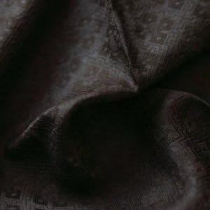 Pode incluir: Close-up de um tecido castanho escuro com um padrão geométrico subtil. O tecido está dobrado, criando sombras e realçando a textura. A trama parece intrincada, sugerindo um têxtil de alta qualidade.