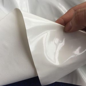 May include: Close-up of white fabric with a shiny, reflective surface. The fabric is being held up, revealing a matte, textured underside. The material appears to be a synthetic textile, possibly for clothing or upholstery.