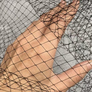 Puede incluir: Primer plano de una red negra con un patrón de diamantes. La red se sostiene, mostrando la textura y transparencia del material. El fondo es gris neutro, resaltando el color oscuro de la red.