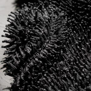 May include: Close-up of a black, textured rug. The rug has a dense, shaggy pile, creating a soft, tactile appearance. The dark color and detailed texture suggest a modern or minimalist design, suitable for home decor.
