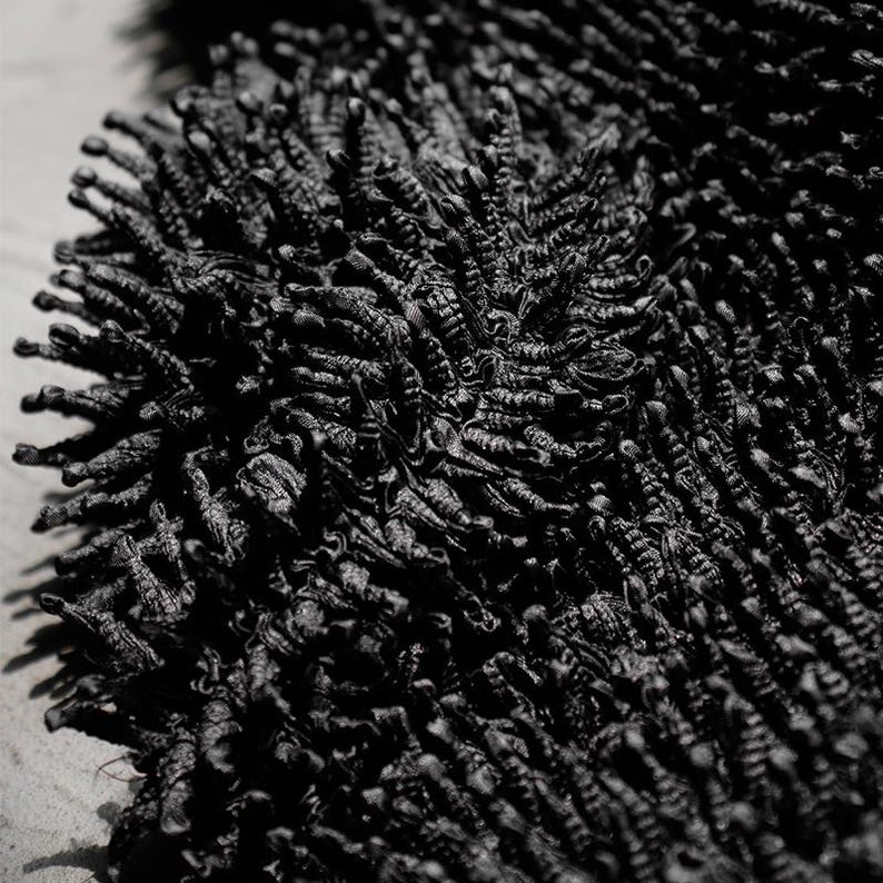 May include: Close-up of a black, textured rug. The rug has a dense, shaggy pile, creating a soft, tactile appearance. The dark color and detailed texture suggest a modern or minimalist design, suitable for home decor.
