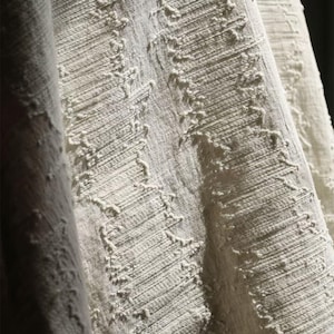 May include: Close-up of a textured, off-white fabric with a woven appearance. The fabric has a slightly distressed look with raised, irregular patterns running diagonally across the surface. The folds and shadows add depth and dimension to the textile.