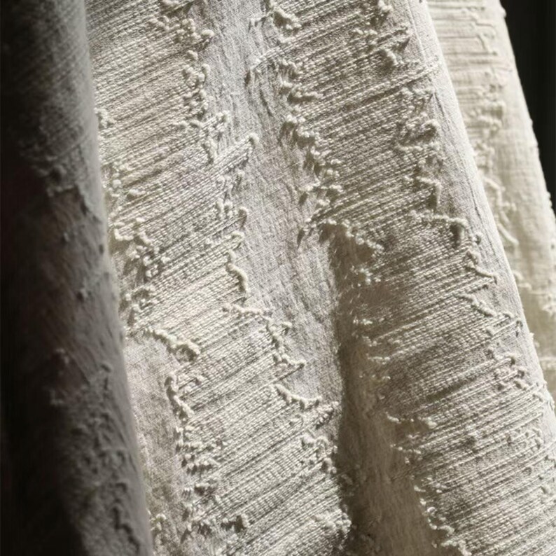 May include: Close-up of a textured, off-white fabric with a woven appearance. The fabric has a slightly distressed look with raised, irregular patterns running diagonally across the surface. The folds and shadows add depth and dimension to the textile.