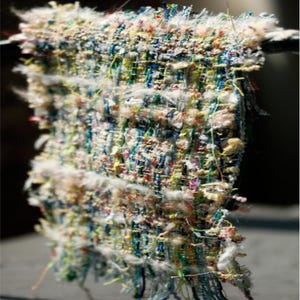 May include: Close-up of a colourful woven fabric sample. The textile features a mix of blue, green, yellow, pink, and white threads, creating a textured, multi-coloured pattern. The edges have a frayed, unfinished look.