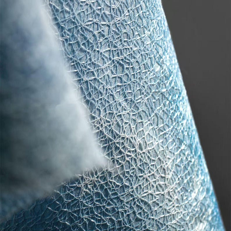 Op de afbeelding: Close-up van een blauwe stof met een witte, webachtige structuur. De stof heeft een driedimensionale uitstraling, waarbij het witte patroon de blauwe basis bedekt. De textuur is ingewikkeld en gedetailleerd, waardoor een visueel interessant oppervlak ontstaat.
